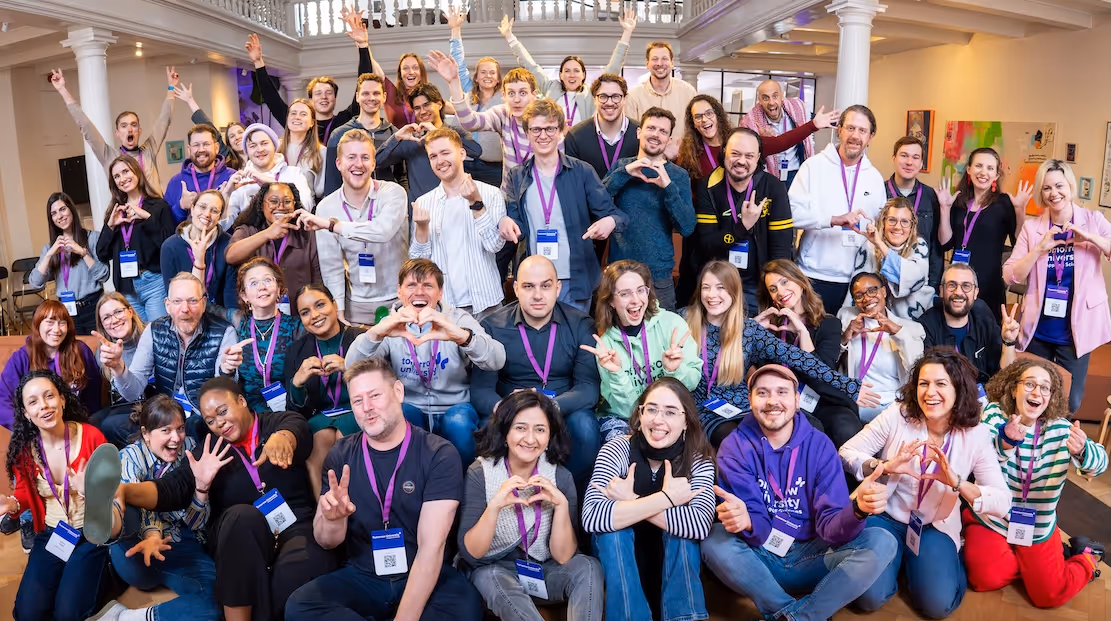 Large diverse group of cheerful people posing indoors, many making heart shapes or peace signs with hands.