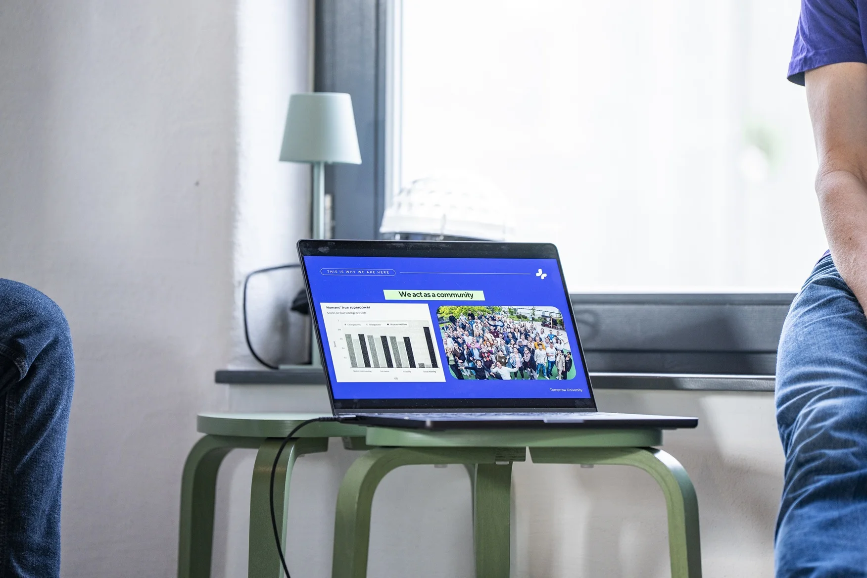 Laptop on a green table displaying a presentation slide with the title 'We act as a community,' including a bar chart titled 'Humans’ true superpower' and a group photo of many people standing outdoors.