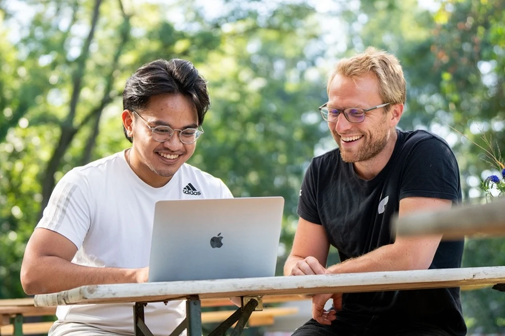 Two men sitting at an outdoor wooden table, smiling and looking at a silver Apple laptop together in a green park setting.