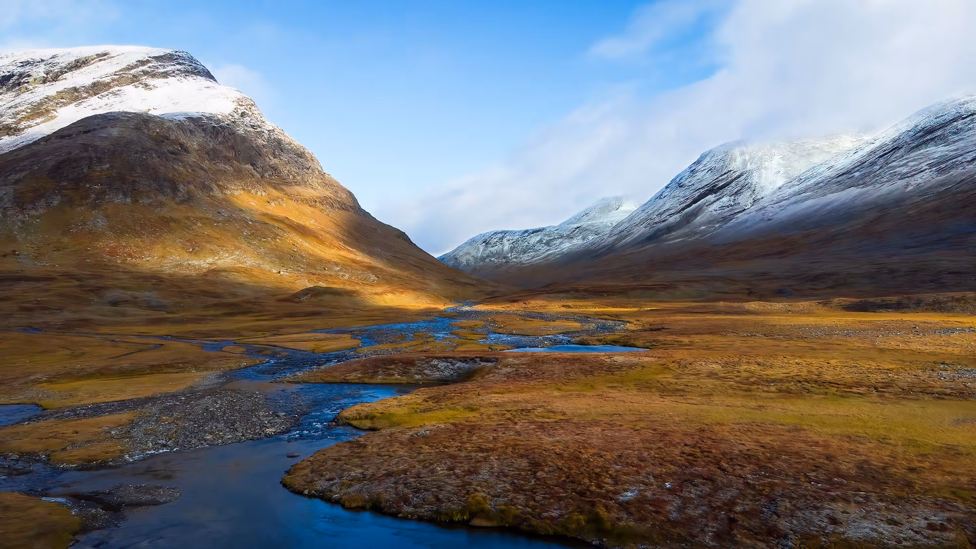 Mjukt ljus över en dalgång med vattendrag och snöklädda berg under en klar himmel.