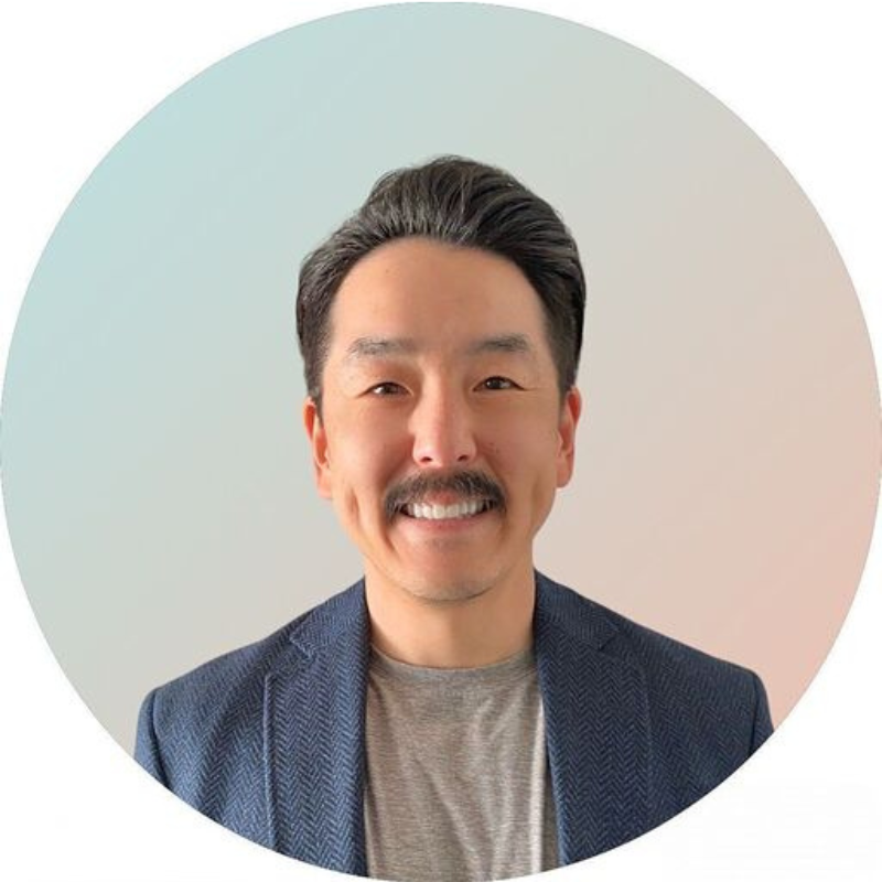 A man wearing a black blazer and light blue shirt, smiling with his arms crossed, framed in a circular border with a light blue outline.