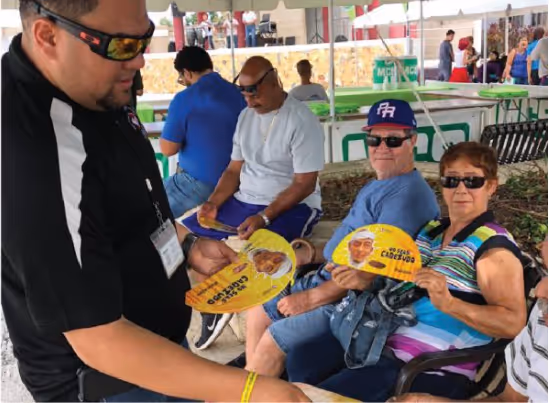 Un hombre con gafas de sol y camiseta negra entrega abanicos promocionales a un grupo de personas sentadas bajo una carpa. Los abanicos tienen la imagen de un candidato político.