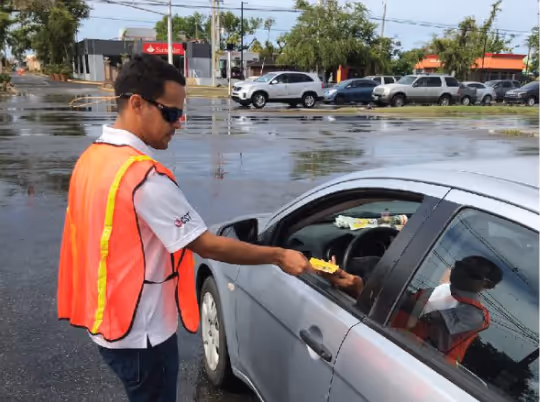 Un hombre con chaleco de seguridad y gafas de sol entrega folletos a un conductor a través de la ventana de su coche en un estacionamiento.