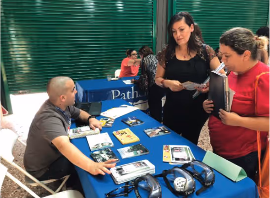 Dos mujeres hablan con un hombre sentado detrás de una mesa cubierta con material informativo. La mesa está en un área al aire libre y tiene varios folletos y dispositivos electrónicos.