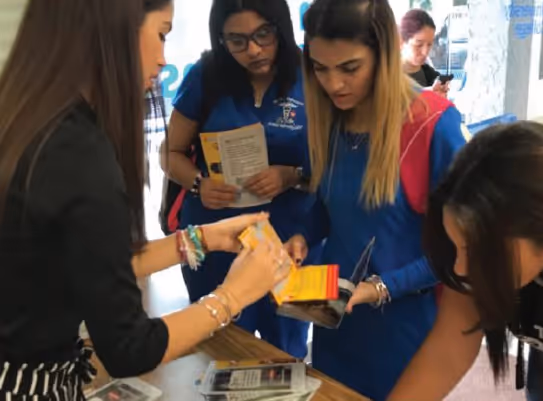Un grupo de mujeres jóvenes, algunas con uniformes médicos, revisan folletos informativos que les entrega otra mujer en un espacio interior.