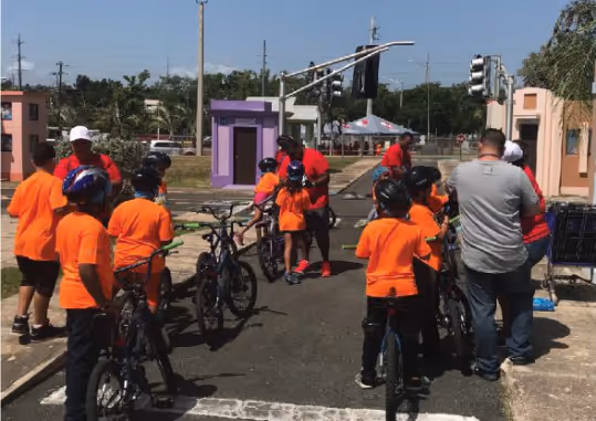 Un grupo de niños en camisetas naranjas y cascos de bicicleta se preparan para una actividad al aire libre. Un adulto supervisa y organiza a los niños mientras están en una calle residencial.