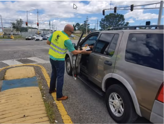 Un hombre con chaleco de seguridad amarillo entrega un folleto a un conductor a través de la ventana de un SUV en una intersección concurrida.