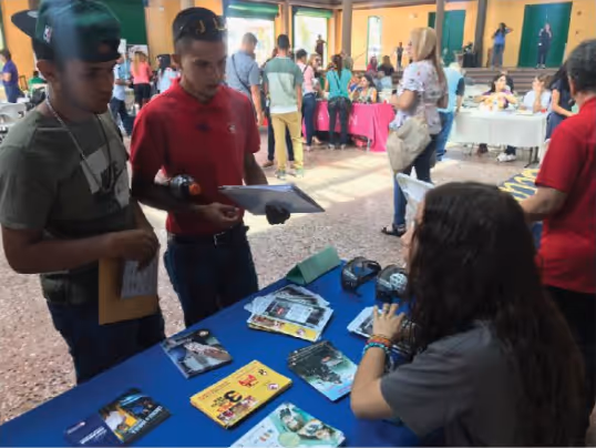 Dos jóvenes conversan con una mujer sentada detrás de una mesa cubierta con material informativo en un evento comunitario. Al fondo, se puede ver a más personas interactuando en diferentes mesas y actividades.
