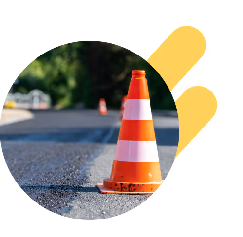 Un cono de tráfico naranja colocado en una carretera recién pavimentada, con más conos visibles en el fondo. La carretera está rodeada de vegetación y árboles verdes.”