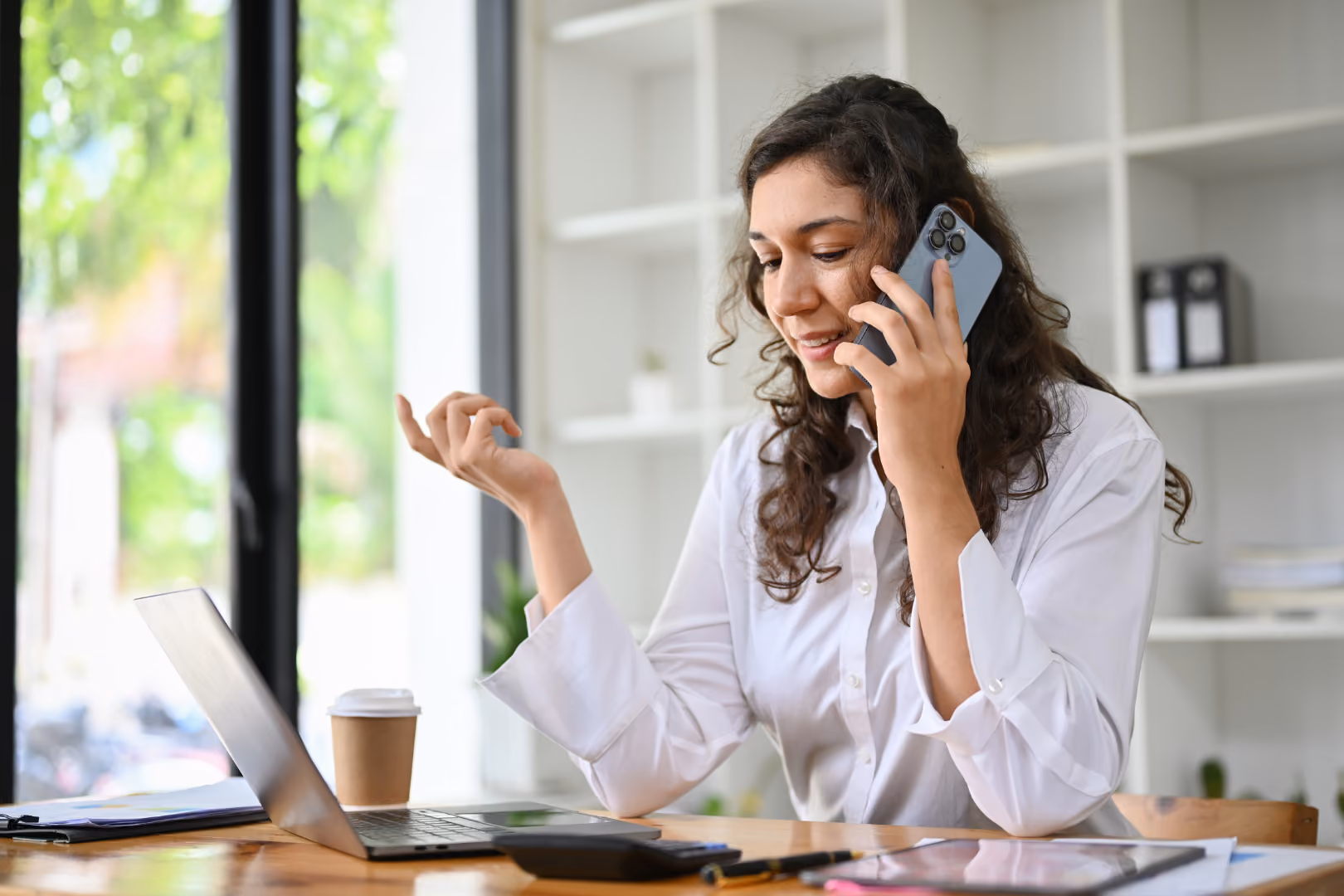 Woman on phone call smiling with laptop in front of her inside her home.