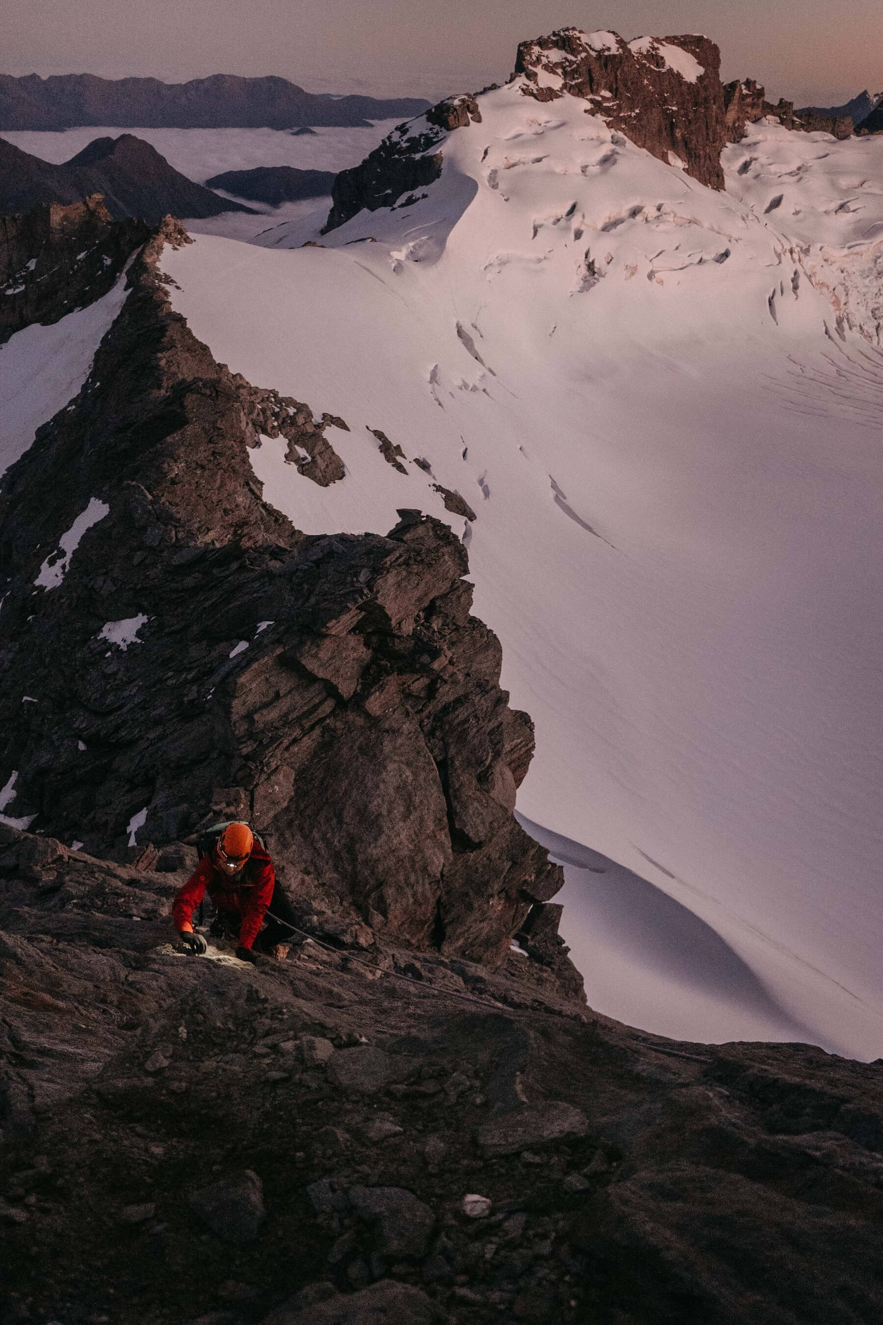 Climber in orange helmet and red jacket ascending rocky mountain with snowy peaks in the background.