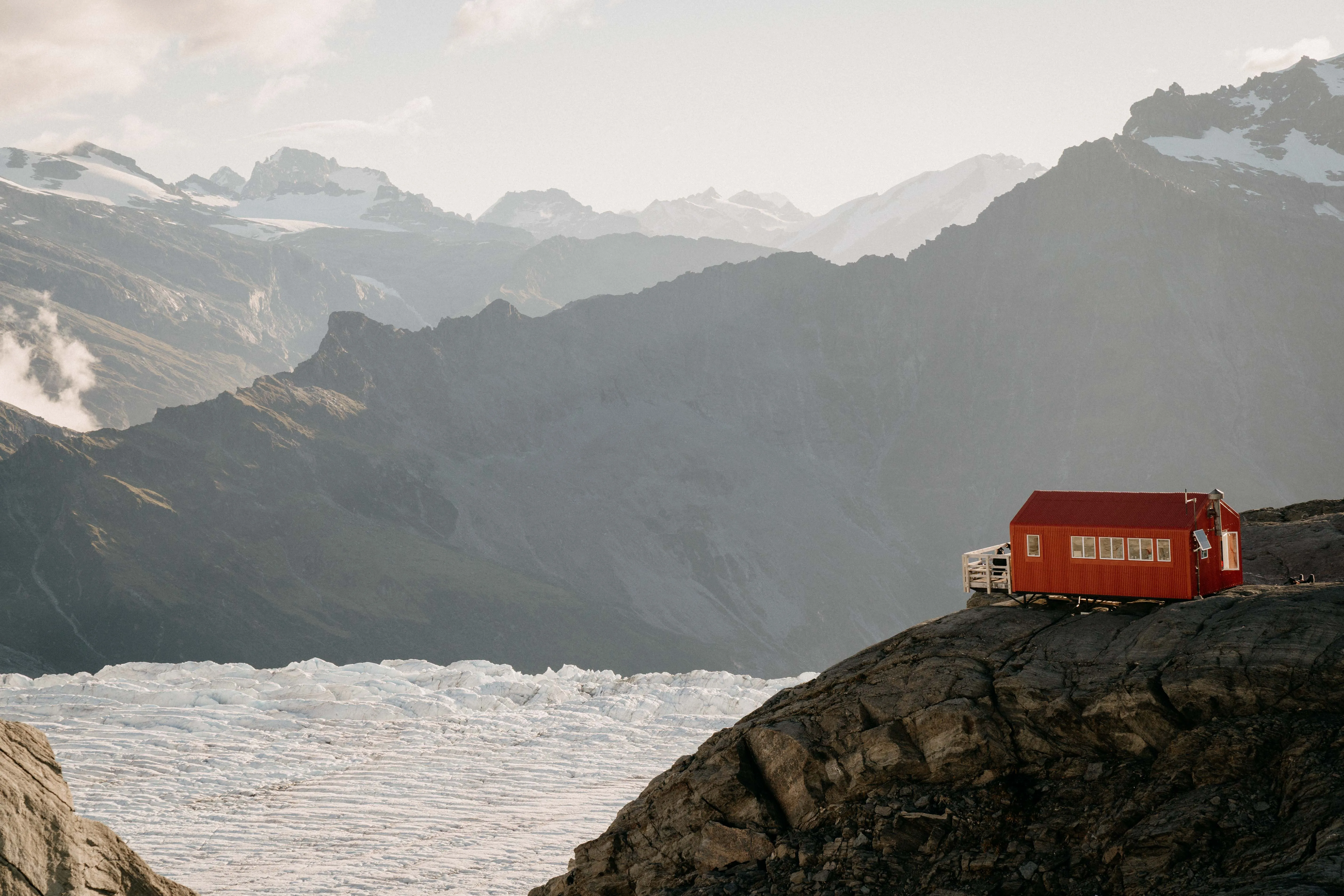 Small red cabin on rocky hillside with glacier below and rugged mountain peaks in the background.