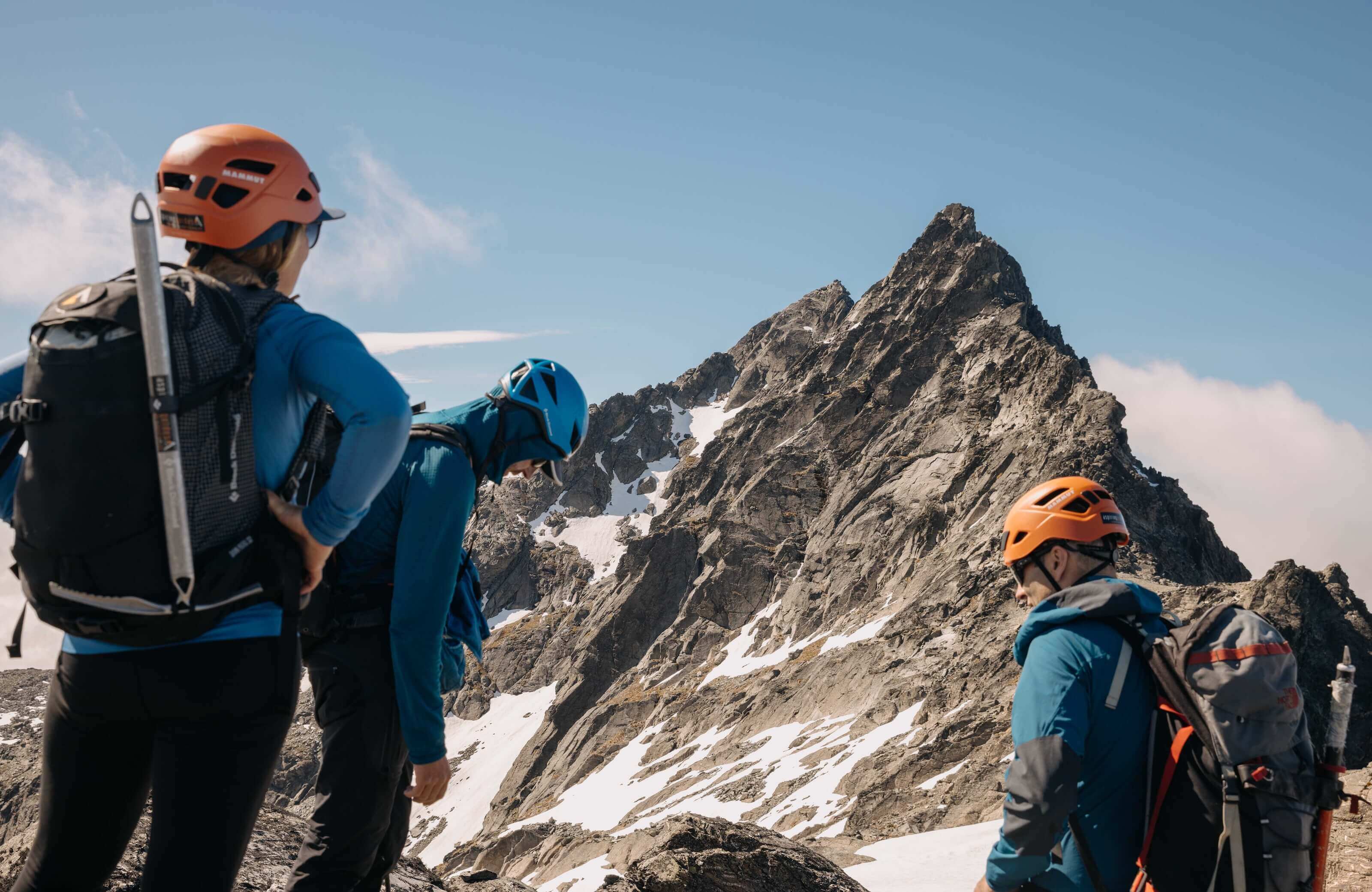 Three climbers in helmets and gear rest near a rocky, snowy mountain peak under a clear sky.
