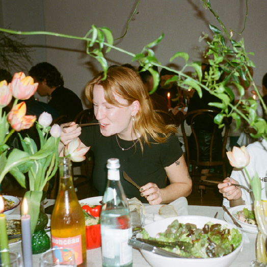 Woman with red hair eating at a dinner table with flowers, bottles, and plates of food.