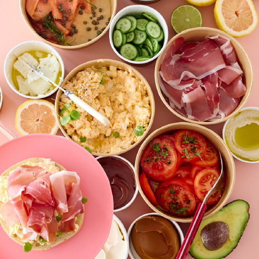 Breakfast spread with scrambled eggs, sliced tomatoes, cucumber, prosciutto, avocado, and various spreads on a pink surface.