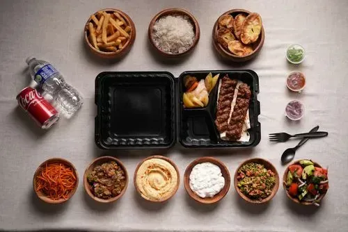 Grilled beef kebabs served with a variety of sides including rice, potato wedges, fries, hummus, tabbouleh, coleslaw, pickled vegetables, and a bottle of water and soda.