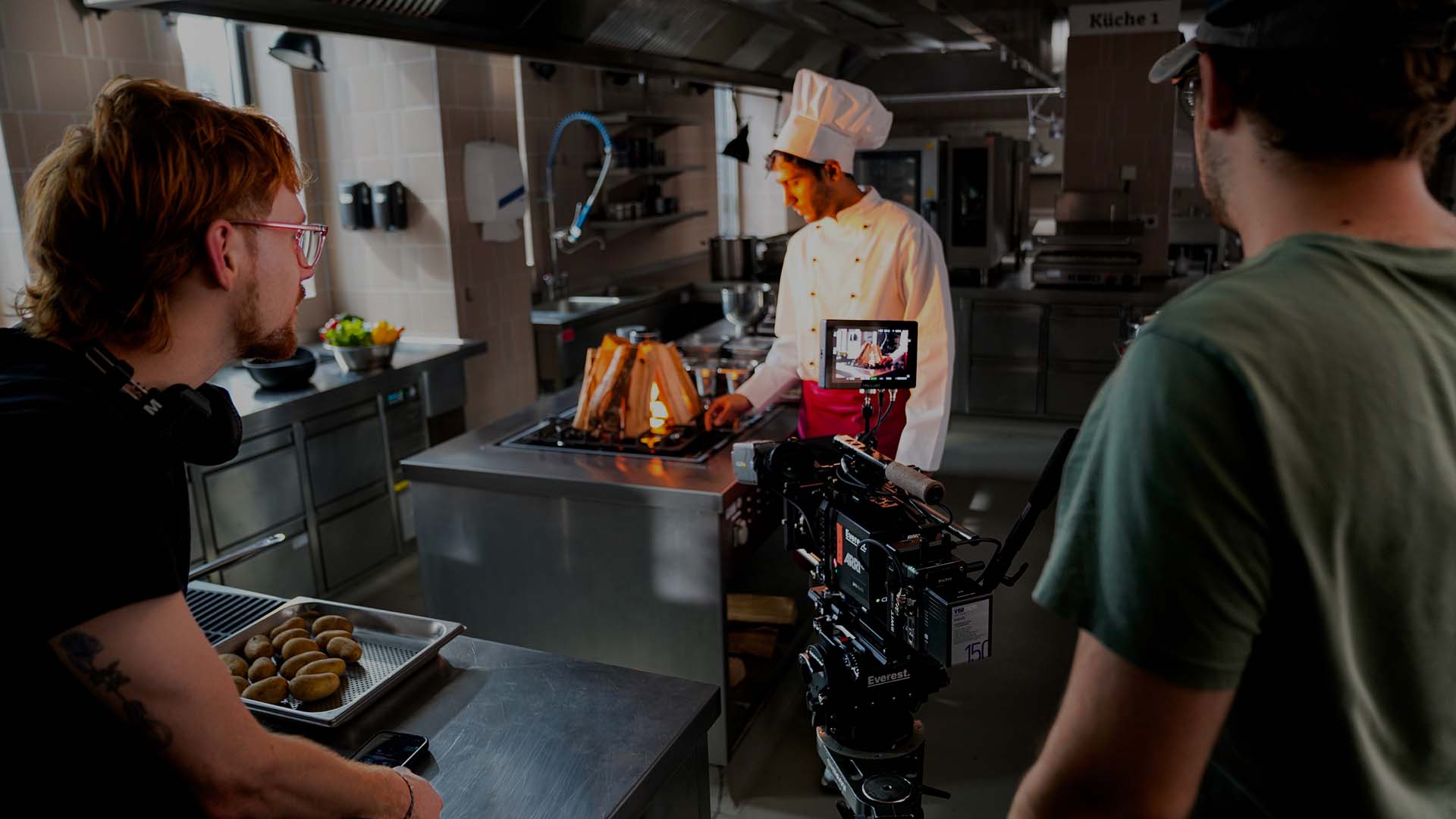 Ein Filmteam dreht in einer professionellen Restaurantküche eine Szene mit einem Koch in weißer Uniform und Kochmütze, der an einem Herd mit offener Flamme arbeitet. Eine Kamera filmt ihn, während zwei Crewmitglieder die Szene beobachten und an der Beleuchtung und dem Ablauf arbeiten.