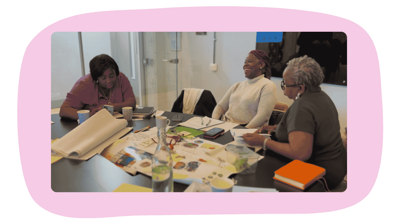 Three women sitting at a table with papers, notebooks, cups, and a water bottle, engaged in discussion.