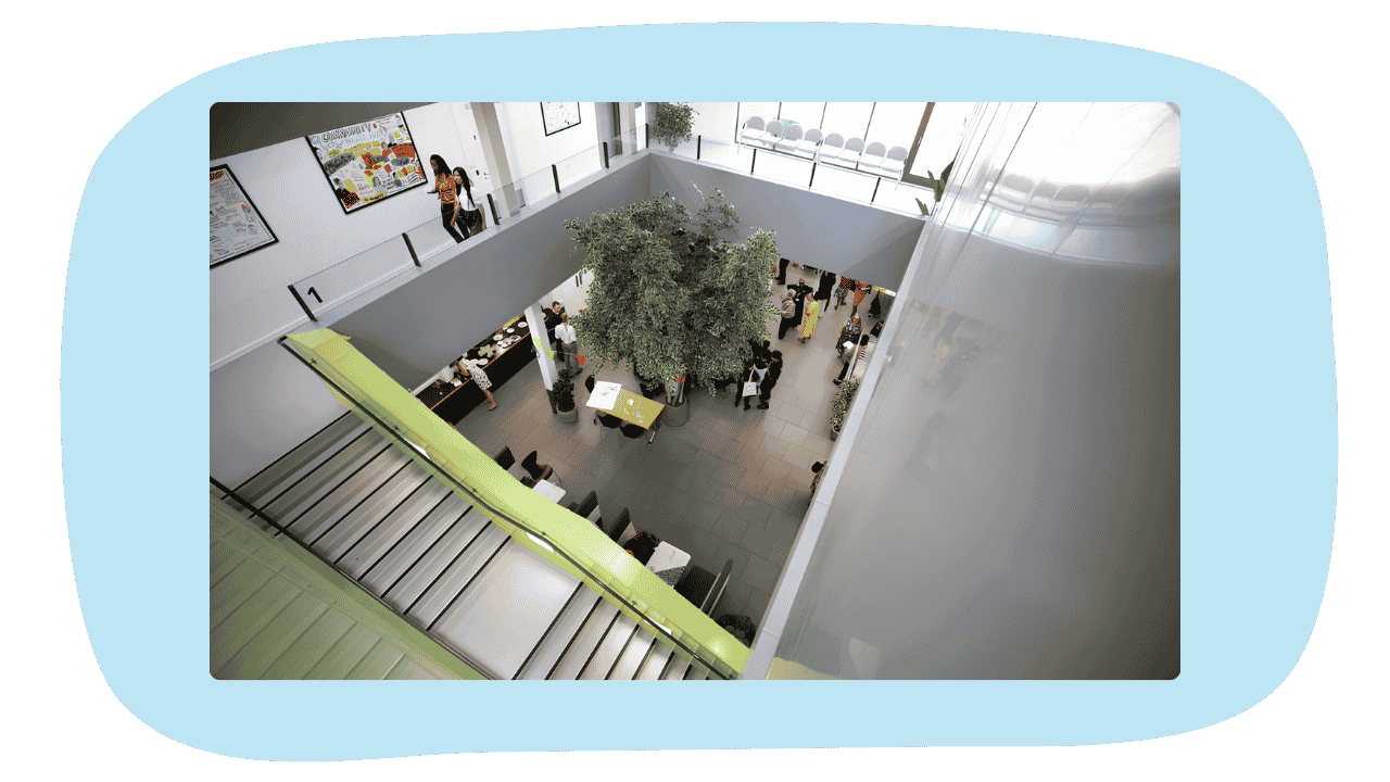 Top view of a modern indoor atrium with stairs, a large tree, and people gathered below.