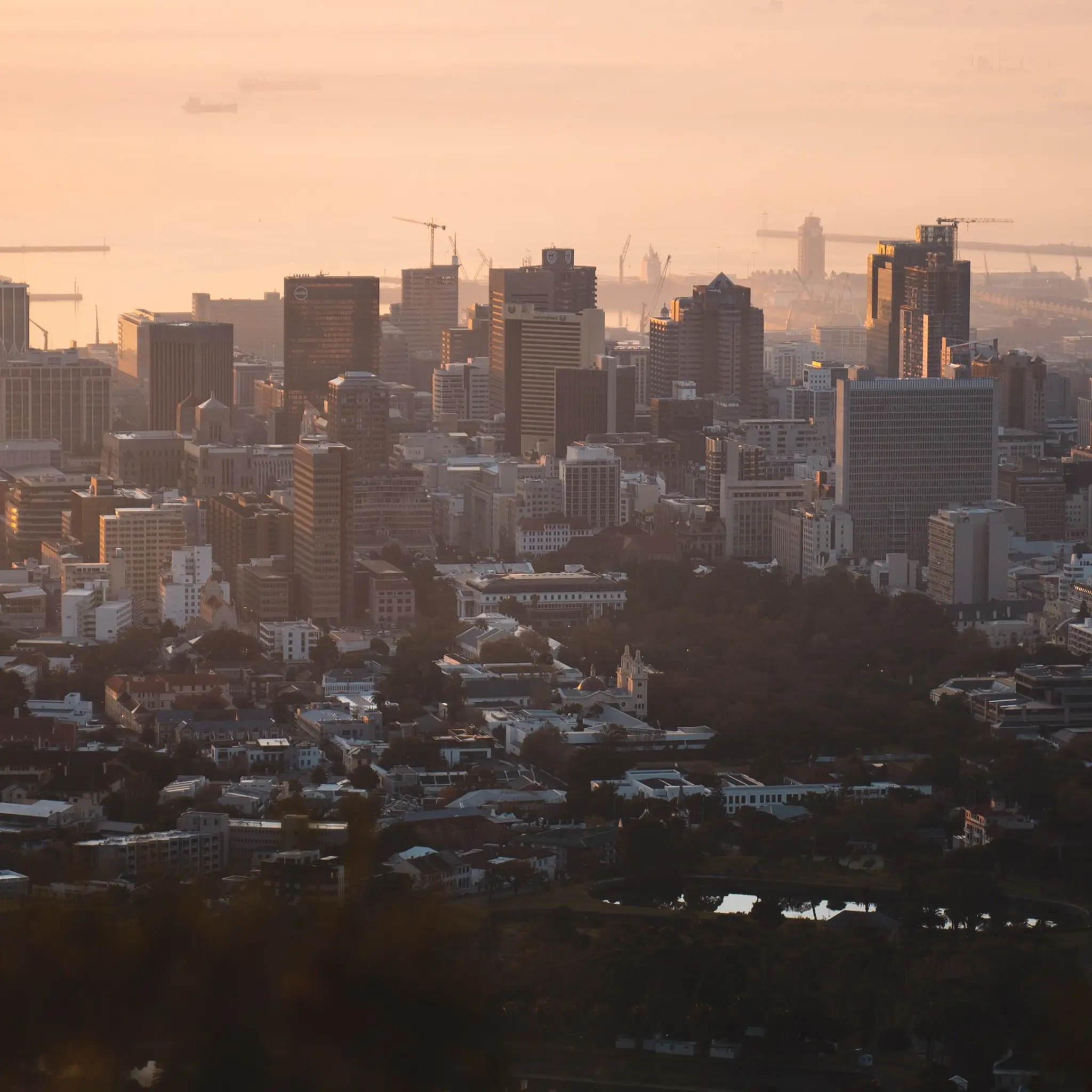 City skyline at sunset with tall buildings, cranes, harbor, and a mix of urban and green areas.