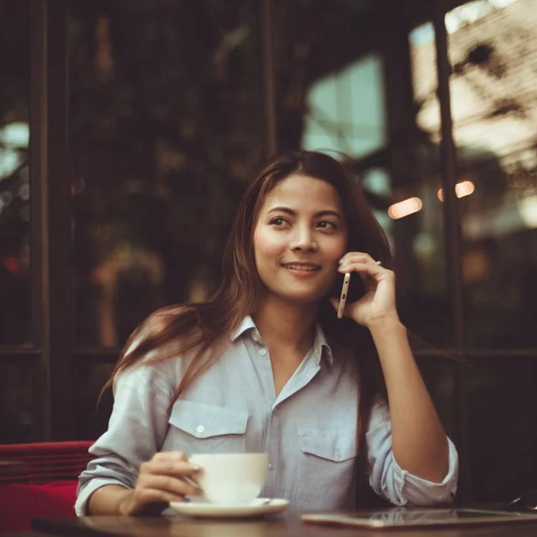 Woman in a light blue shirt holding a cup and talking on a phone at a cafe table.