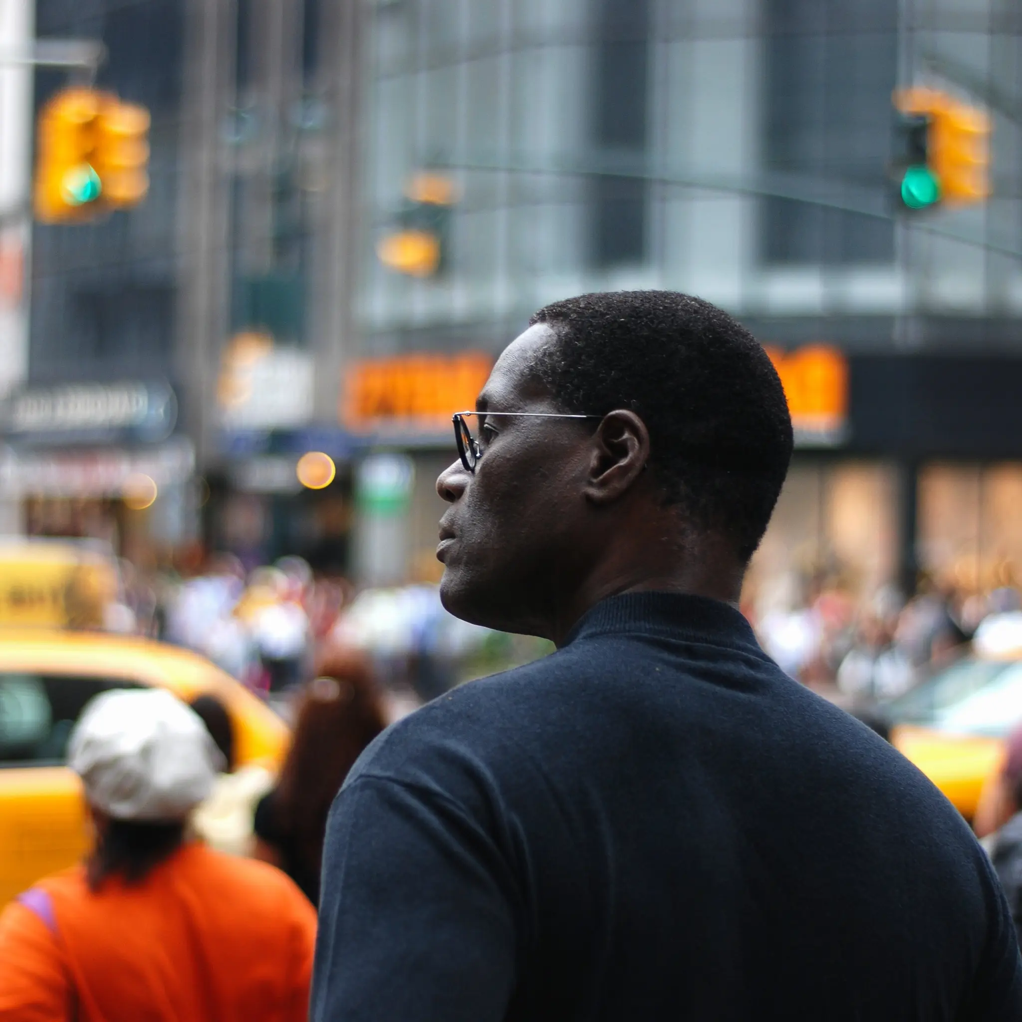 Man in glasses and dark shirt standing on busy city street with blurred traffic lights and crowd.