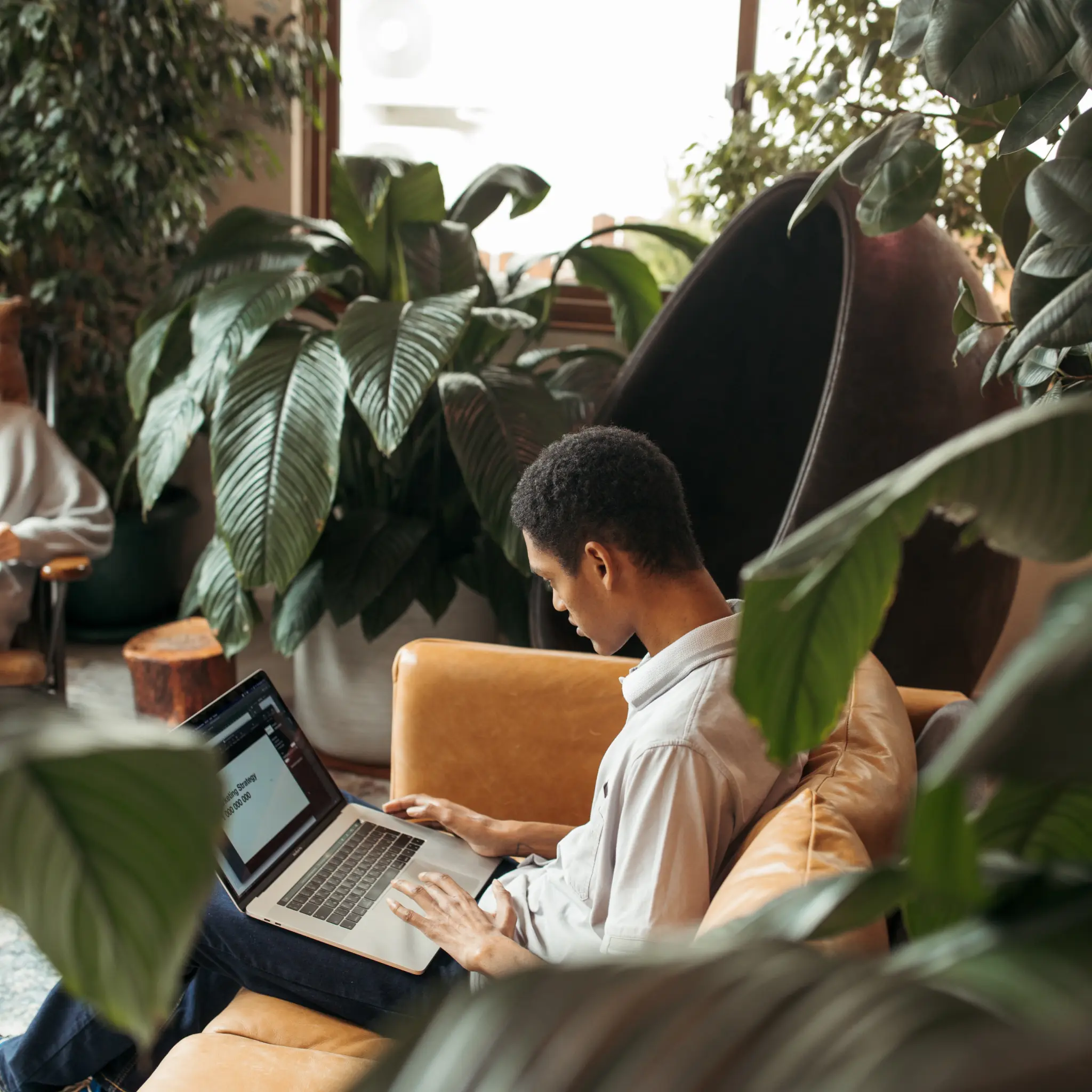 Man sitting on a tan leather couch working on a laptop in a plant-filled room.