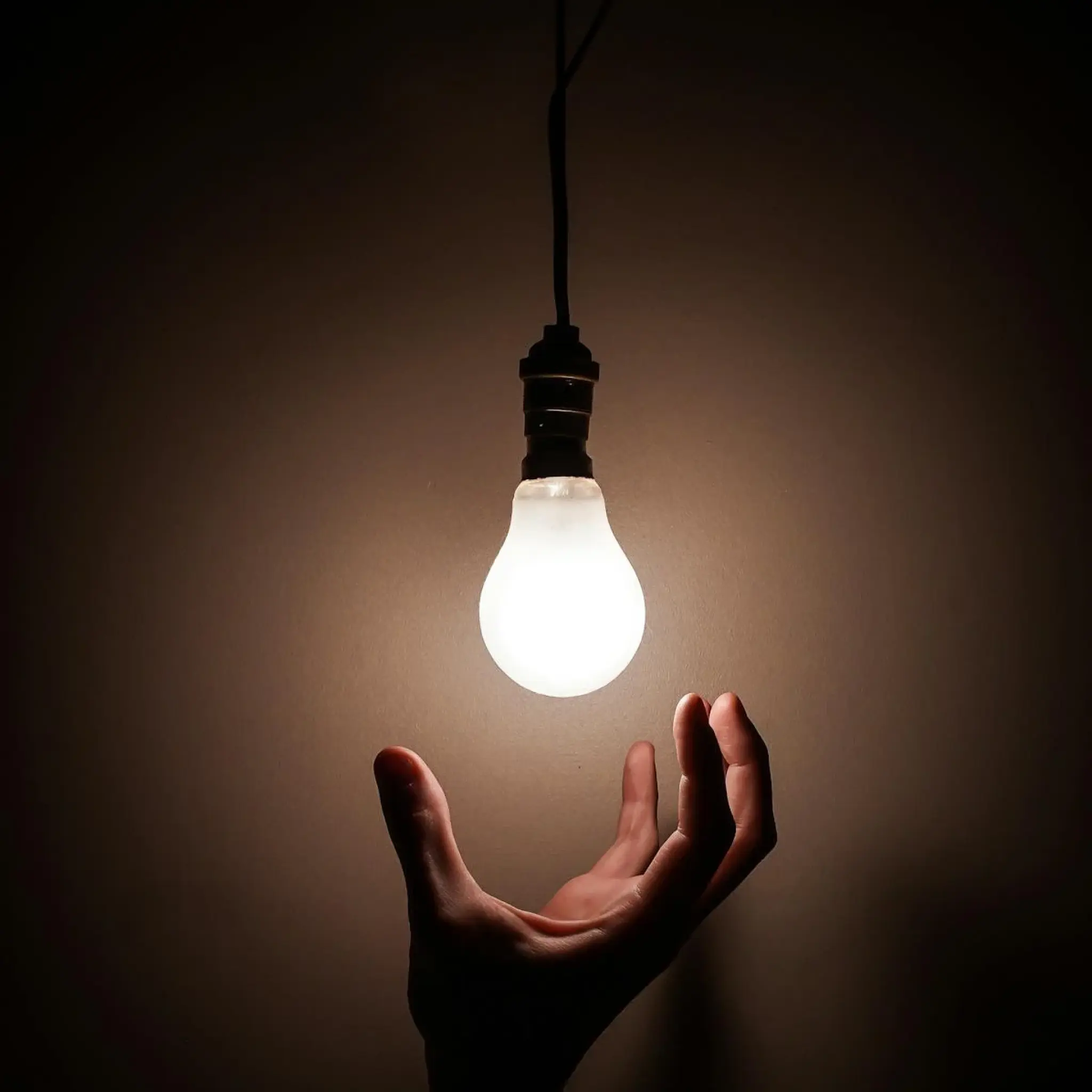 Hand reaching towards a glowing light bulb hanging from a cord against a dark background.