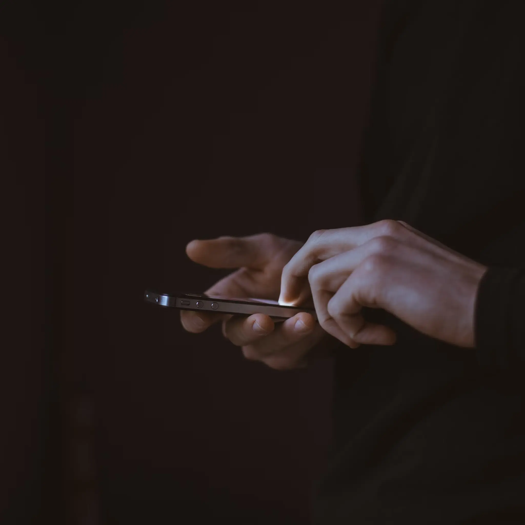 Person using a smartphone with one hand, tapping the screen with the other hand in low light.