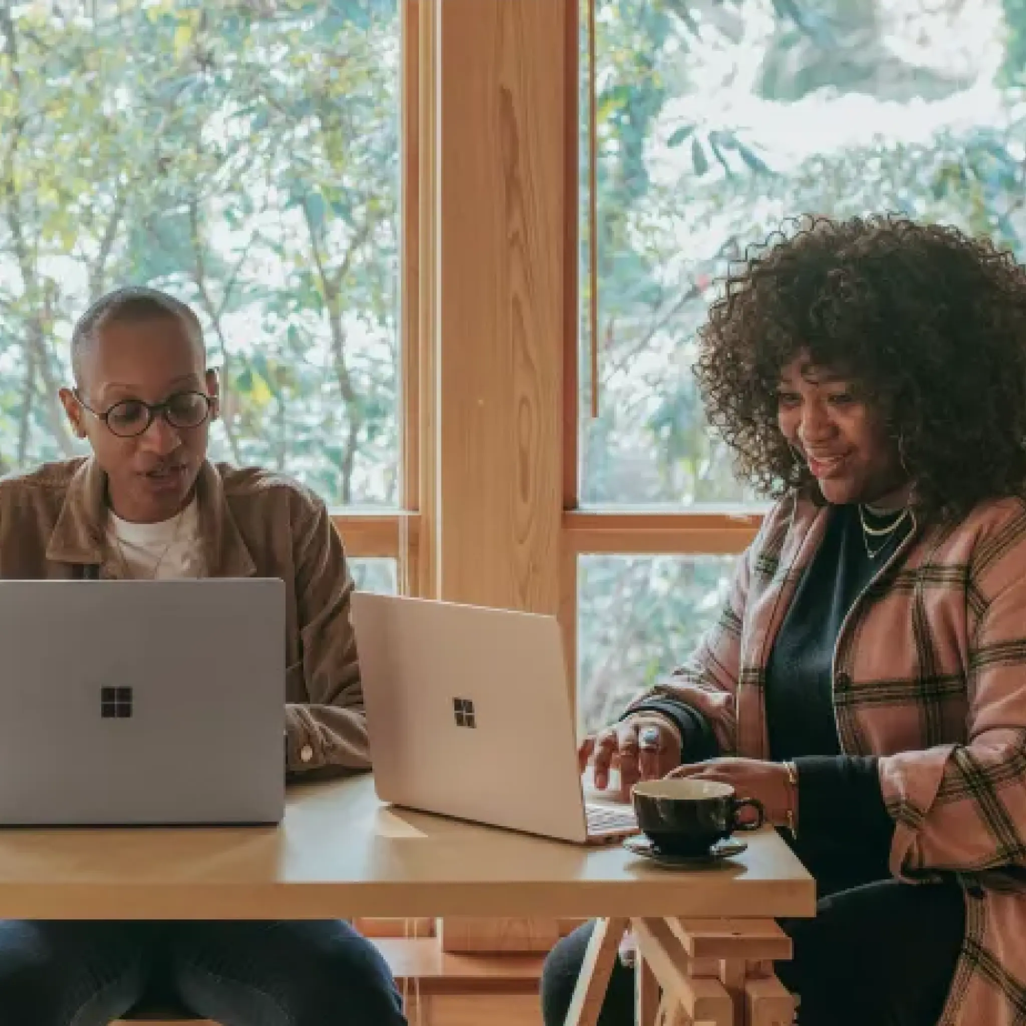 Two people sitting at a wooden table working on laptops with large windows and greenery outside.