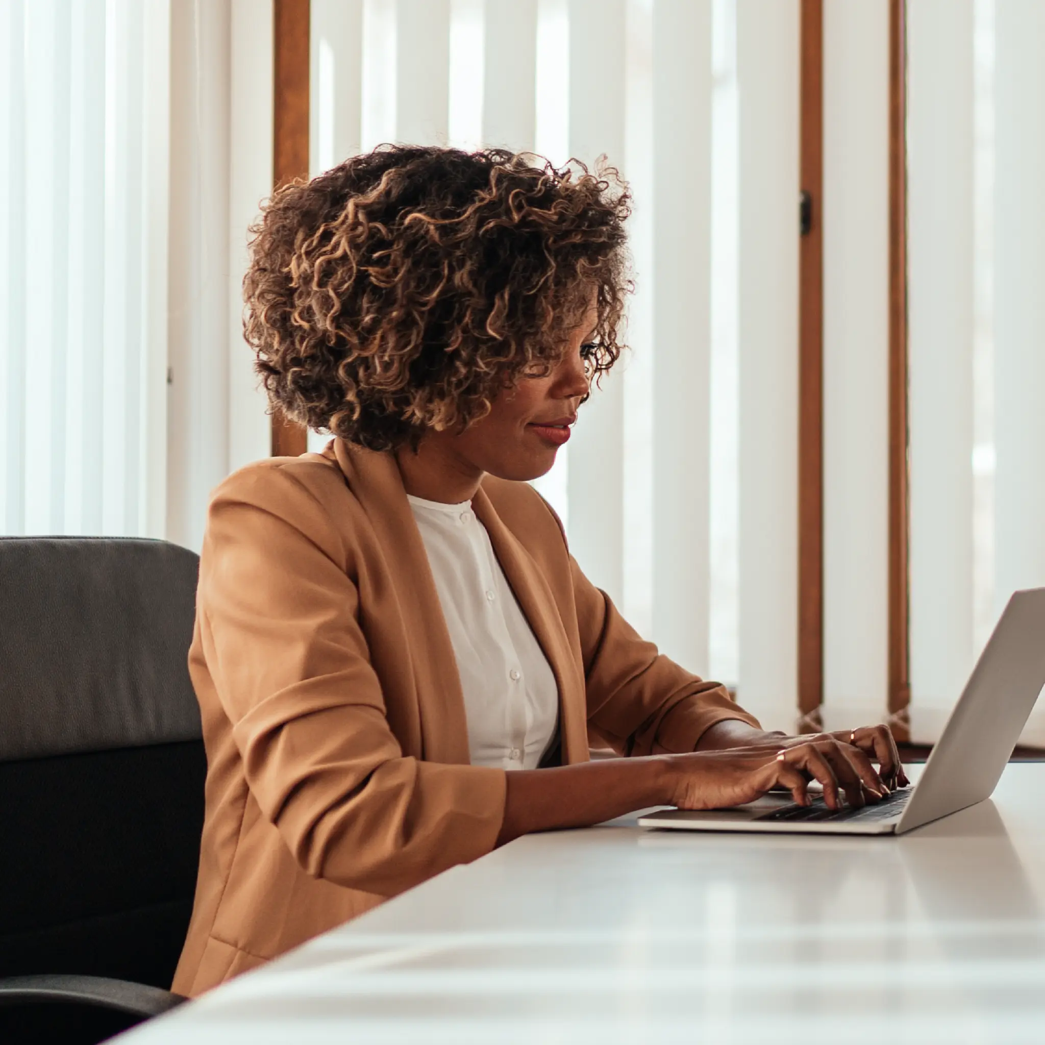 Woman with curly hair in a tan blazer typing on a laptop at a white desk in an office.