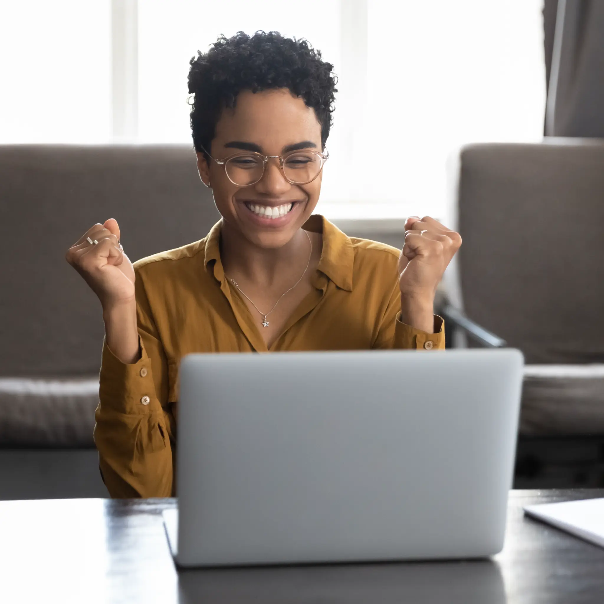 Smiling woman with glasses in brown shirt shows excitement sitting at a desk with a laptop.