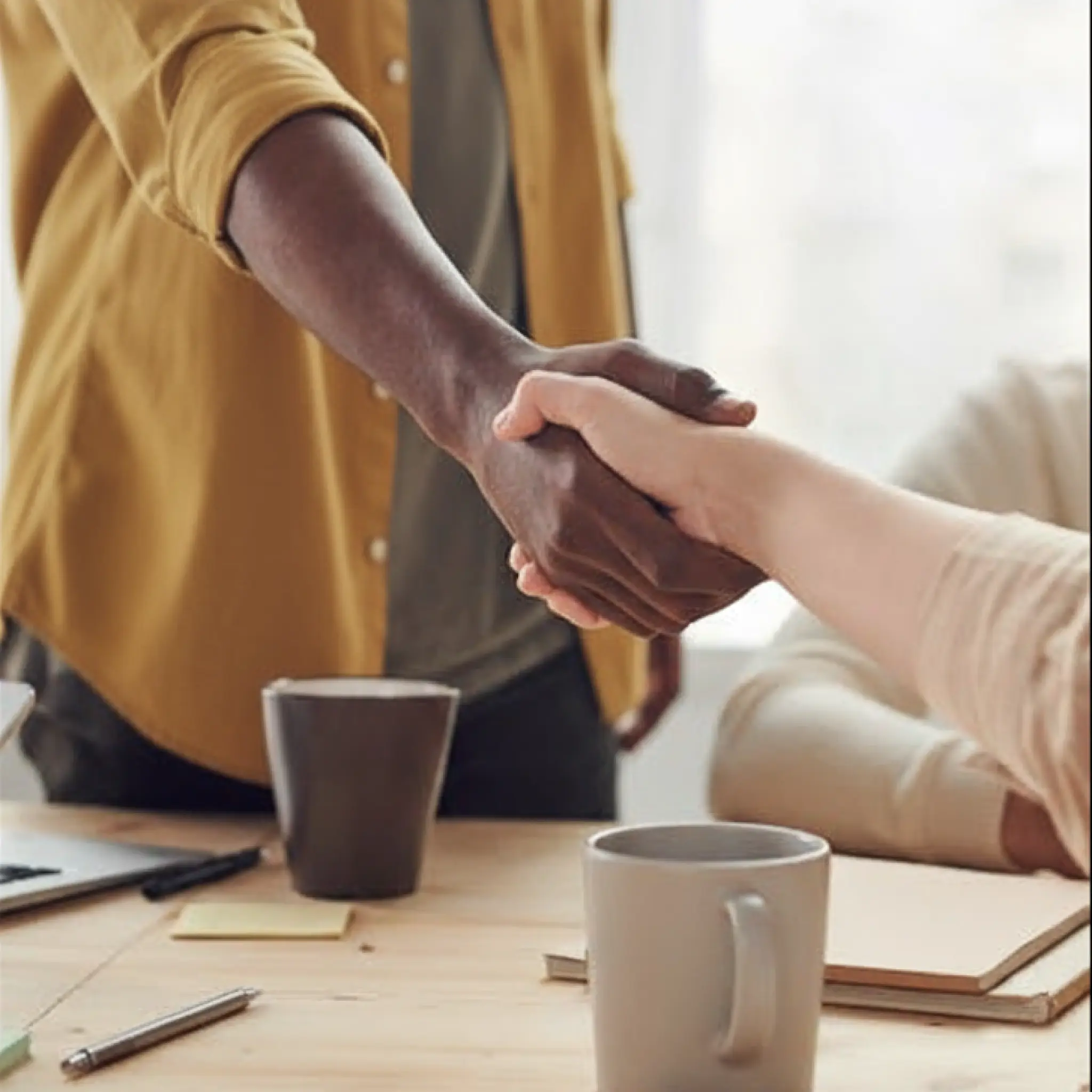 Two people shaking hands over a wooden desk with mugs, notebooks, and pens.
