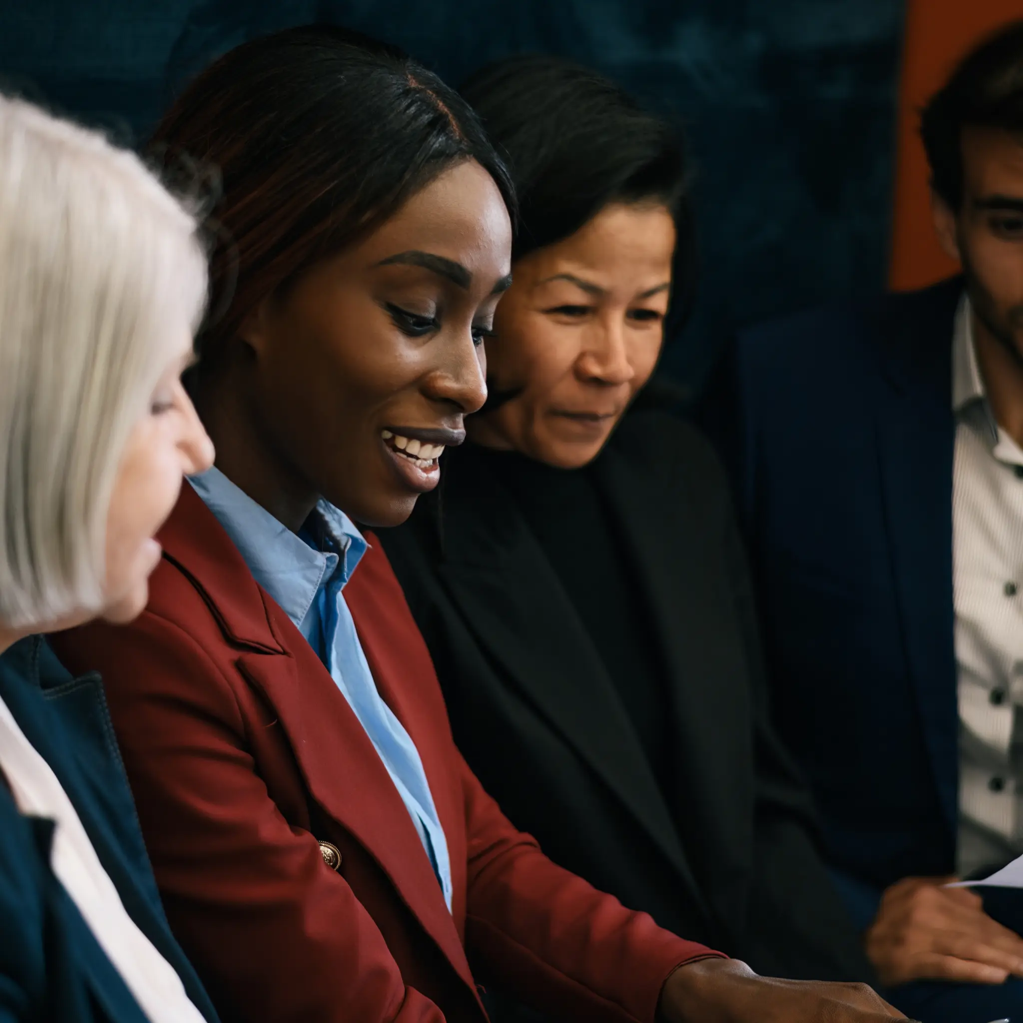 Four diverse professionals closely engaged in a discussion, with a focus on a smiling Black woman in red blazer.