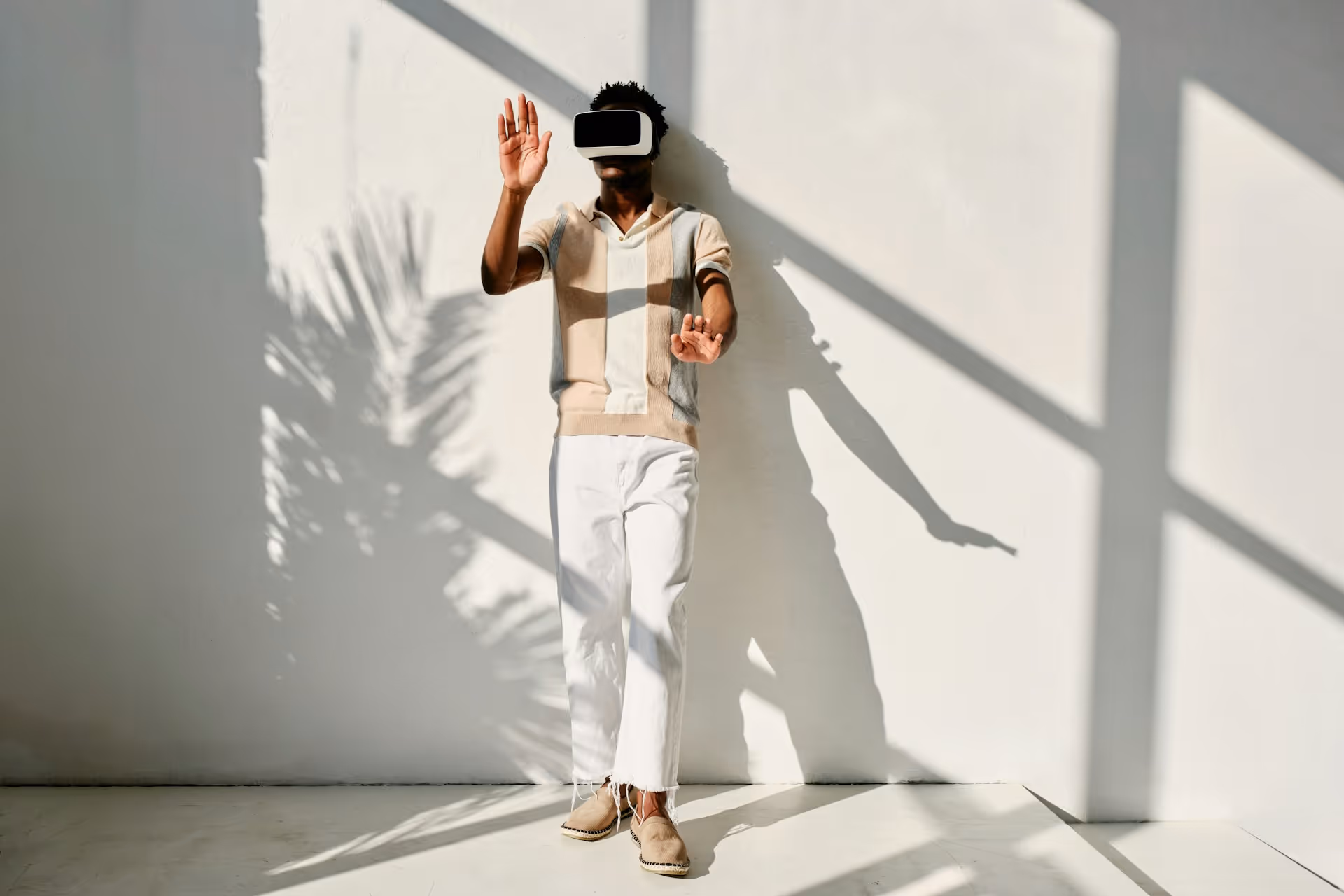 Man wearing a virtual-reality headset, standing against a white wall with shadows and raising his hands.