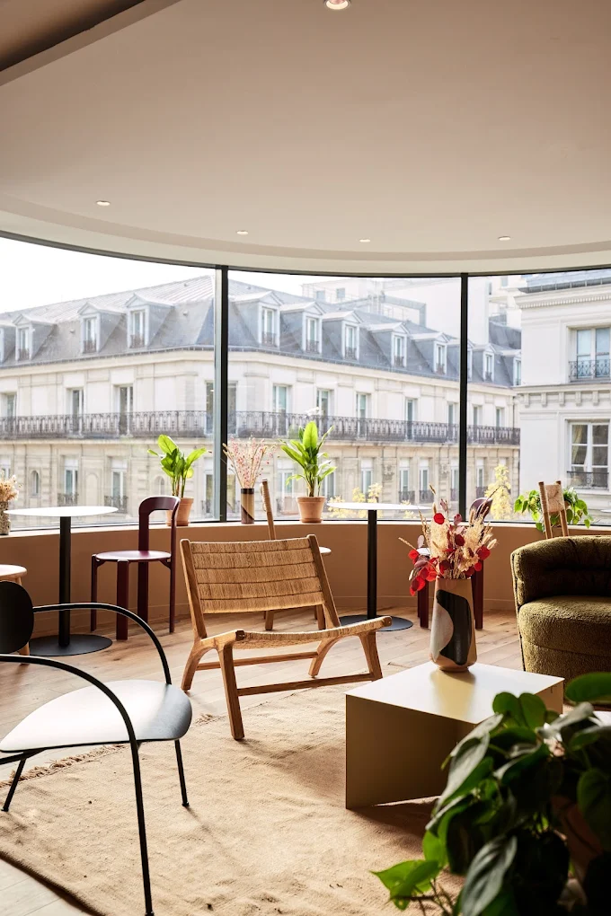 Modern lounge with armchairs, a coffee table and plants in front of a large window overlooking historic buildings.