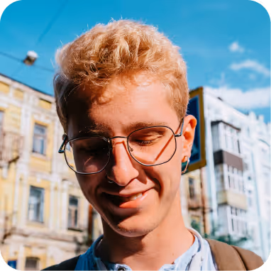 Young person with short curly blond hair and round glasses smiling outdoors with buildings and blue sky in the background.