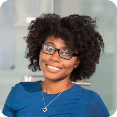 Smiling woman with curly hair and glasses wearing a blue top and a heart-shaped pendant necklace.