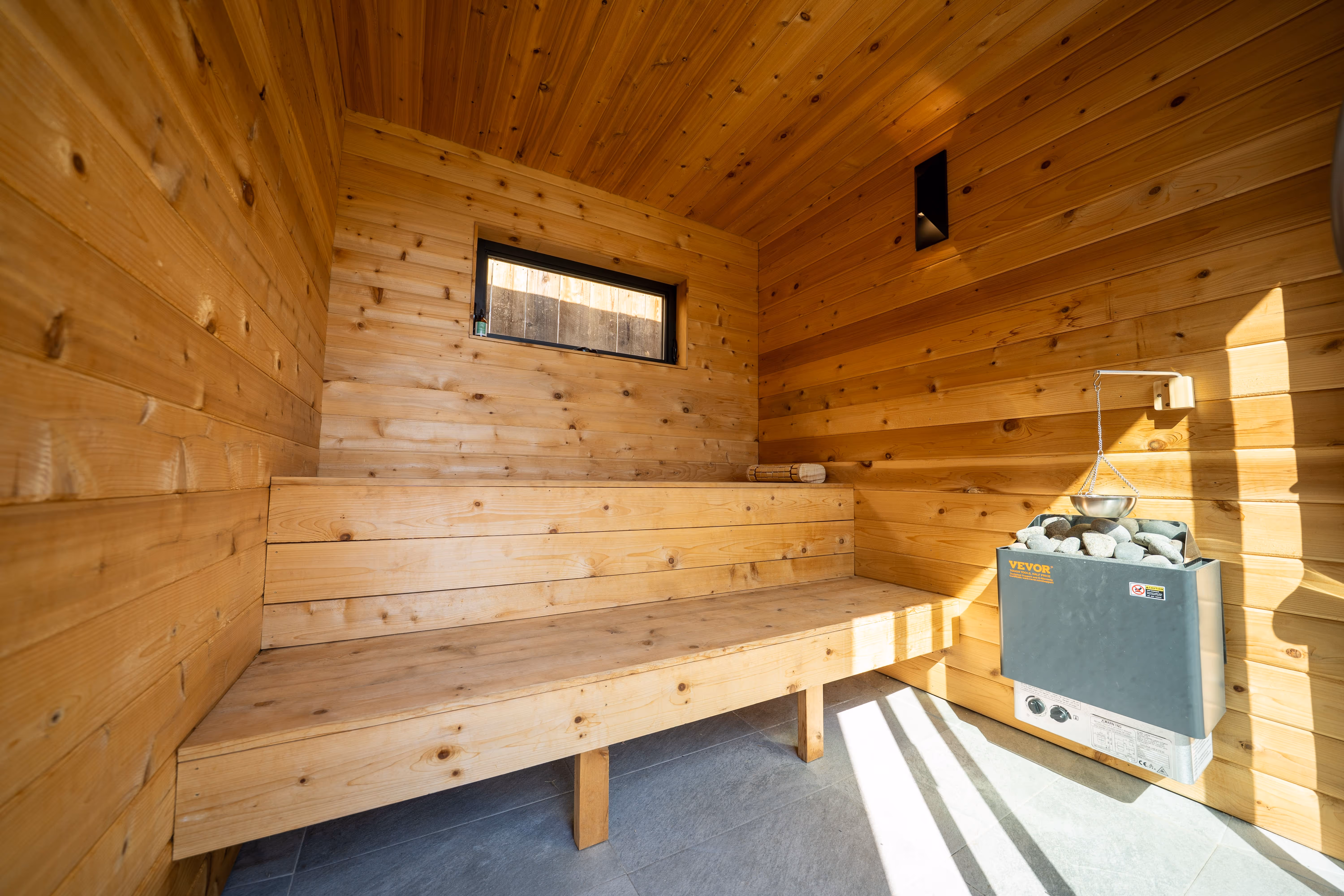 Interior of a wooden sauna with bench seating, a small window, and a Vevor heater with sauna stones and water ladle.