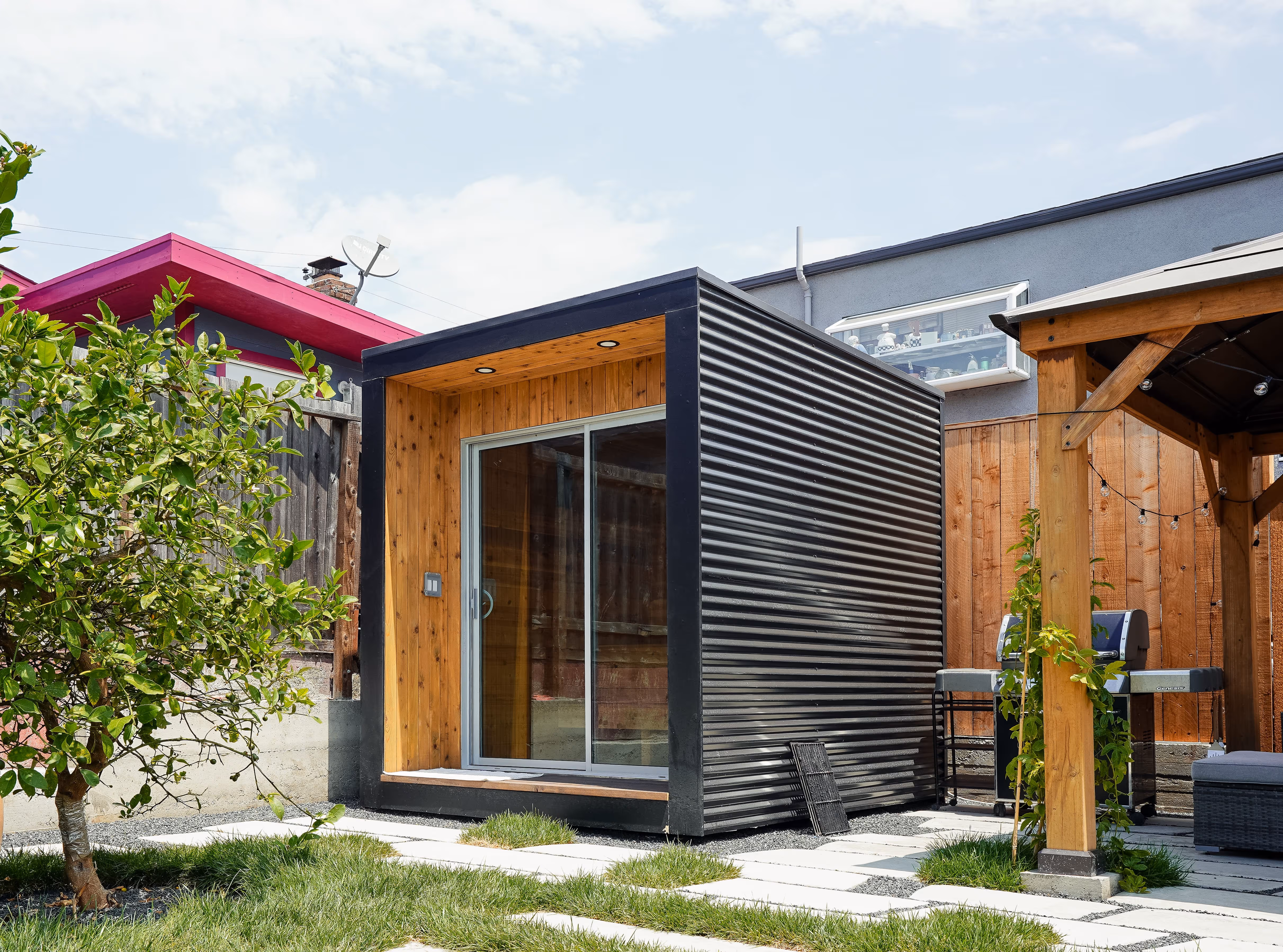 Modern small backyard sauna with black corrugated metal exterior and wooden interior, sliding glass door, grass, and a wooden pergola.