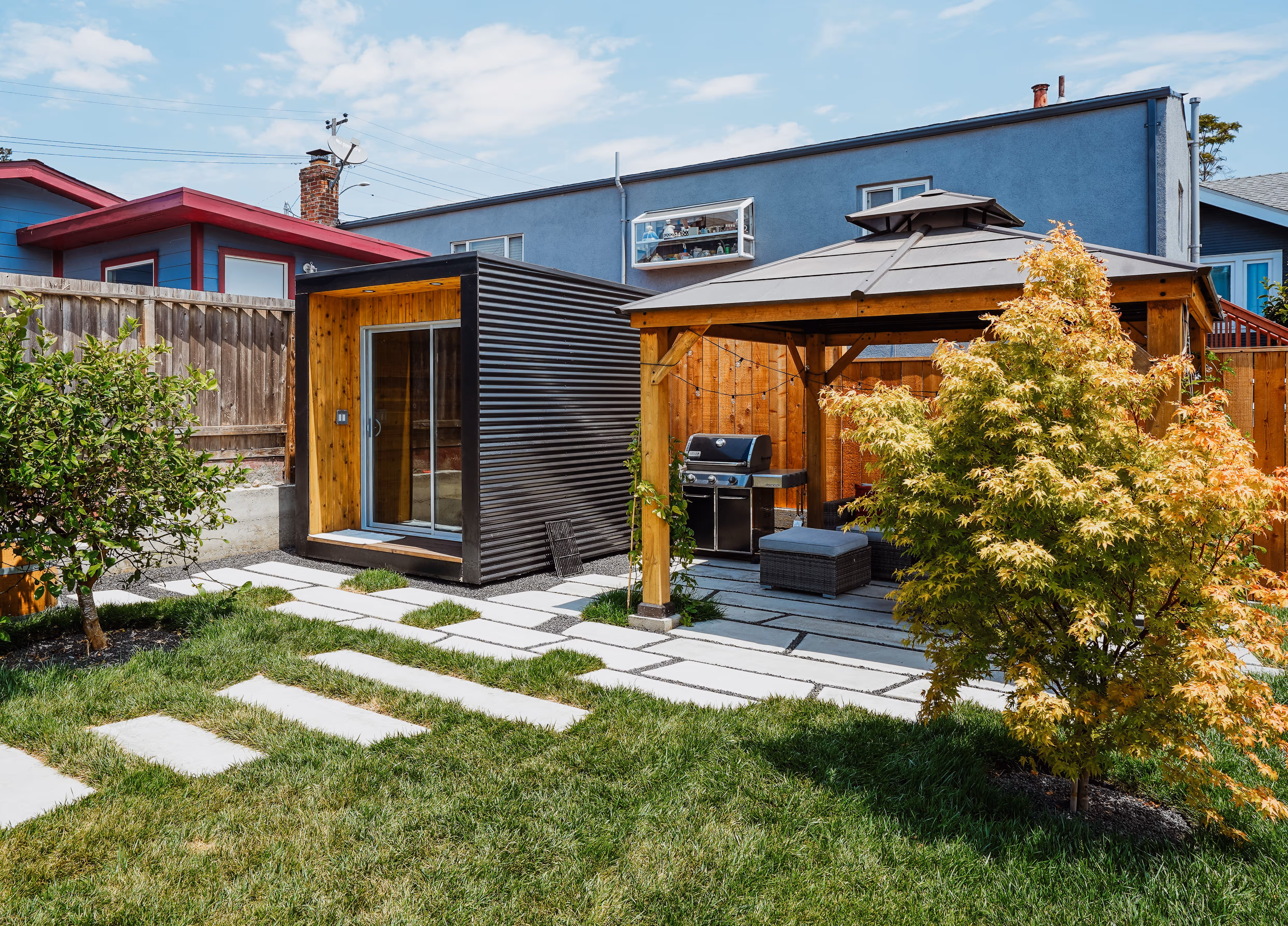 Backyard with green lawn, small trees, stone pathways, a black sauna with wooden interior, and a wooden pergola with a barbecue grill and outdoor seating.