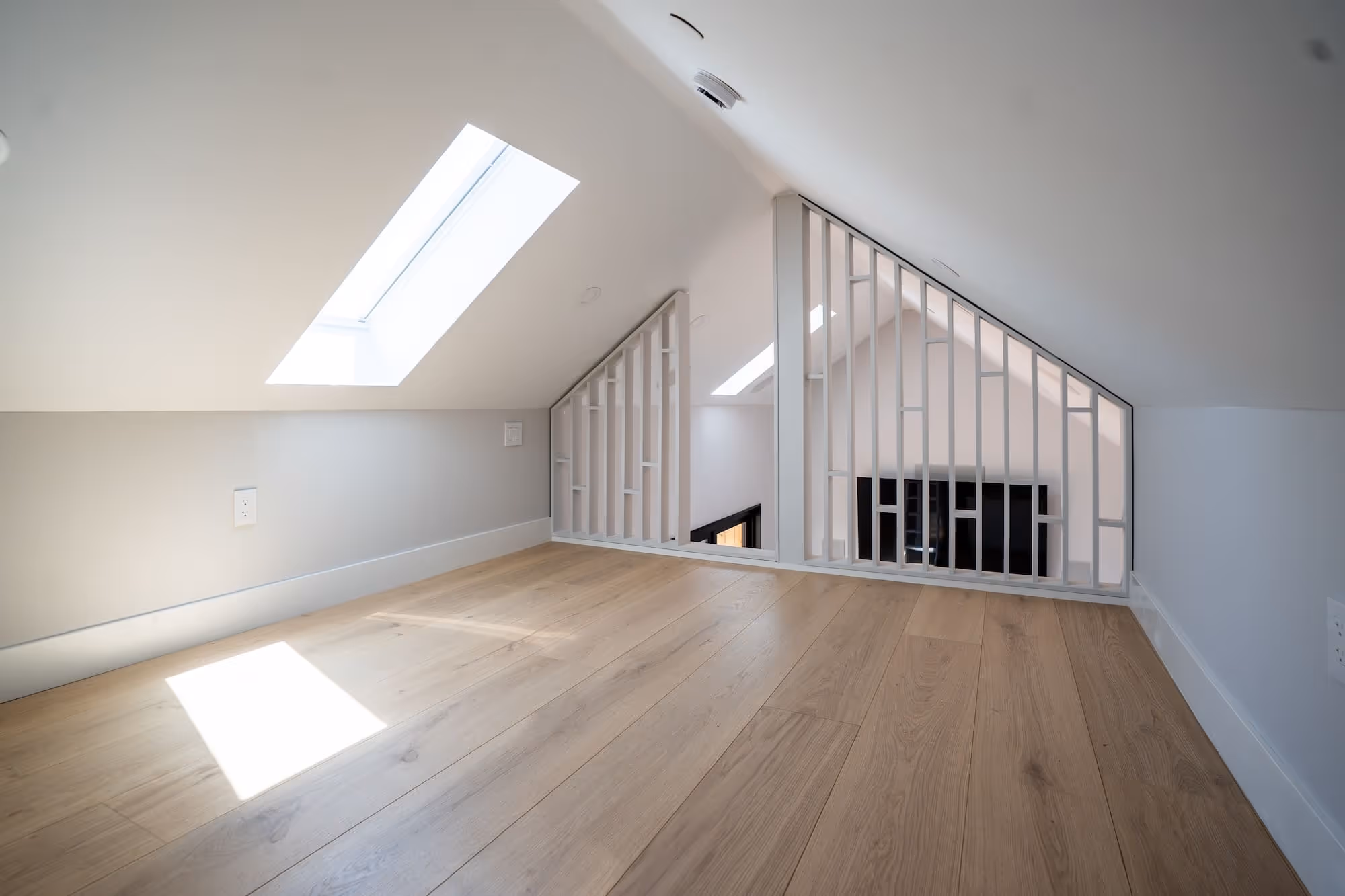 Bright attic space with wooden flooring, white walls, skylight window, and white vertical railing overlooking a lower floor of an ADU.
