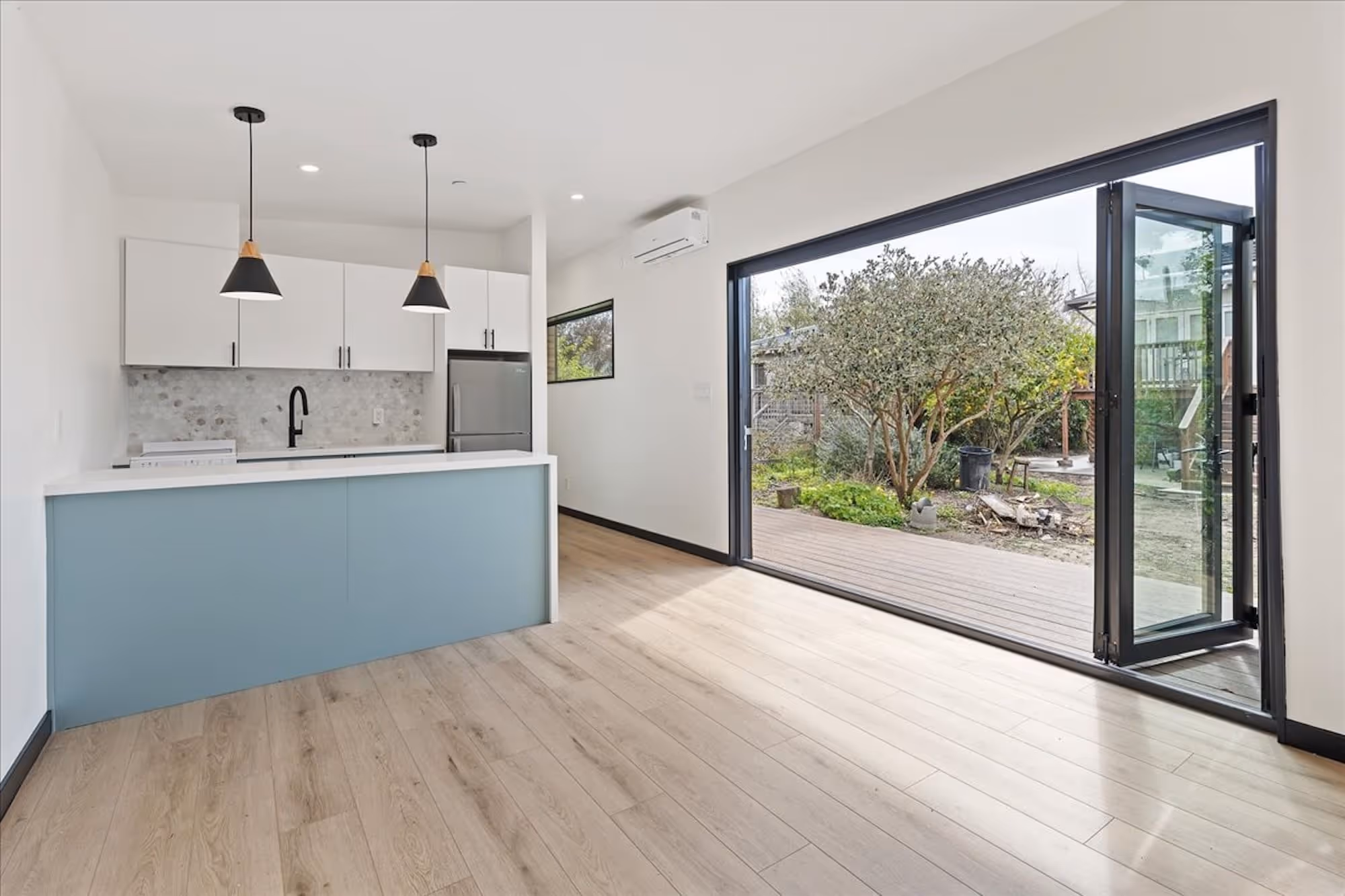 Interior of an ADU featuring a modern kitchen with light wood flooring, blue island, white cabinets, pendant lights, and large folding glass doors opening to a backyard.