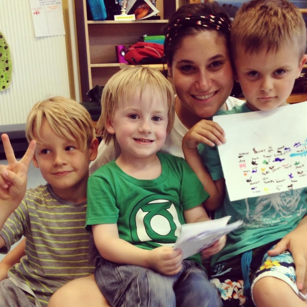Friendly babysitter smiling at the camera while sitting on the floor with three young children holding artwork