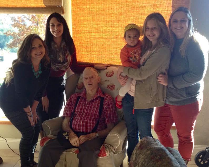 A group of six people, including a man sitting down and five women standing around him, posing for a photo indoors.