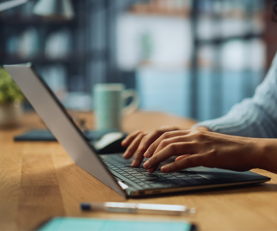 Person sat at a desk typing at a laptop. They can only be seen from the shoulders down but they are wearing a light blue jumper and they have shoulder-length brown hair.