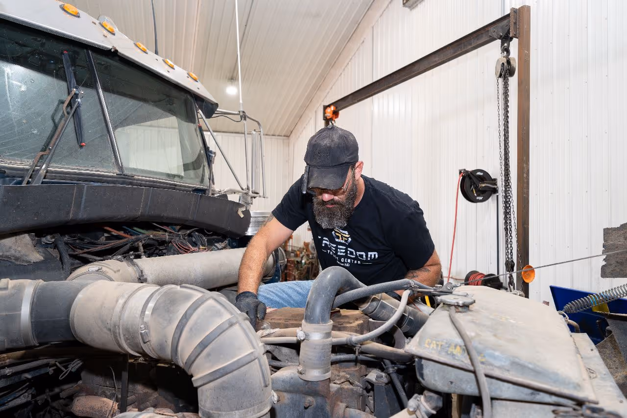 Technician working on a diesel engine inside a clean, well-lit repair shop, wearing a Freedom Truck Center shirt and protective gloves.