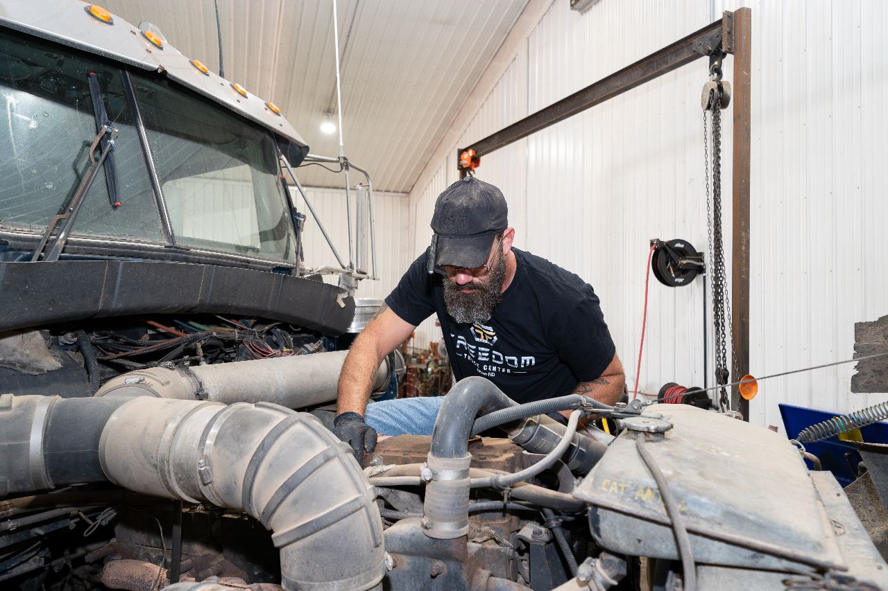 Technician working on a diesel engine inside a clean, well-lit repair shop, wearing a Freedom Truck Center shirt and protective gloves.