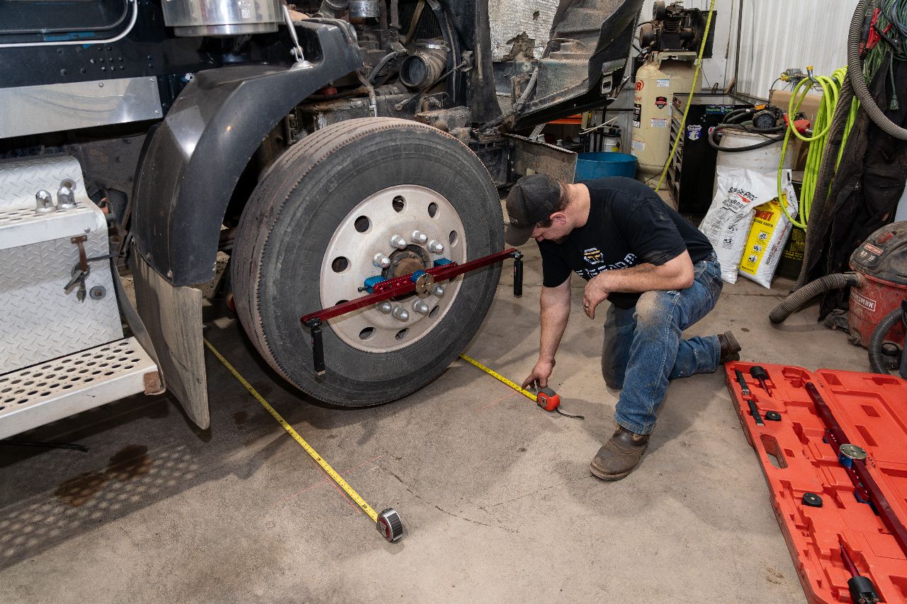 Mechanic measuring truck wheel alignment using specialized tools and tape measures inside a repair shop with equipment and toolboxes visible.