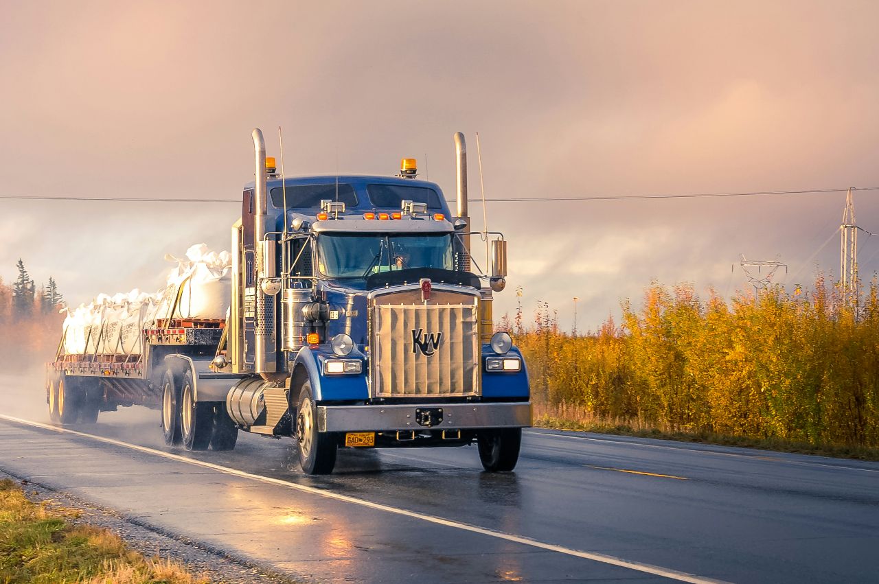 Blue Kenworth semi truck hauls covered flatbed load on wet highway, kicking up mist under cloudy sunset sky.